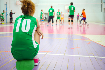 Girl in green sits on ball in gym during volleyball game, back view, playing people out of focus