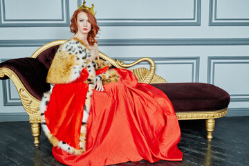 Red-haired woman in red dress, cloak and with crown on head sits on couch