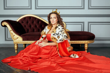 Fototapeta premium Woman in red dress, cloak and with crown on head sits on floor near couch in room