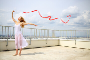 Young woman with blond hair in white standing with red curly ribbon on the roof of a multistory...