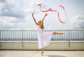 Pretty gymnast in white with raised hands with red curly ribbon standing on a leg on the roof of a multistory building