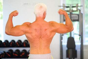 Fototapeta na wymiar Middle-aged instructor shows his muscles in gym, back view, shallow dof