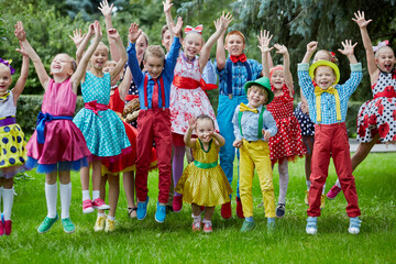 Group portrait of fifteen children dressed in bright dance suits jumping at grassy lawn with hands...