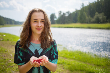 Happy girl teenager holds red wild strawberry under rain near river at summer day