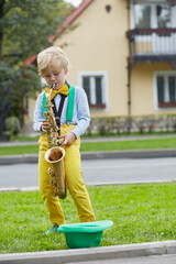 Little boy in dancing suit plays saxophone on grassy lawn against two-storied house near road, hat...