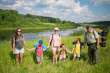 Three adults and three children stand with backpacks near river at sunny day during hike