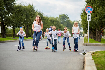 Woman and five children on scooters, roller skates and skateboard ride on street