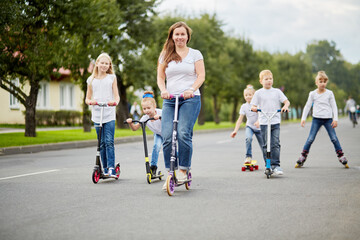 Woman and five children on scooters, roller skates and skateboard ride on street 