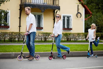 Man, woman and girl ride on scooters in front of house on street