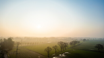 The sun's gentle rise diffuses through mist, casting a hazy glow over a quiet rural expanse dotted...