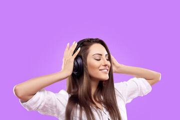 Beautiful woman smiling and enjoying favorite music, via wireless headphones over purple background. Girl uses wireless earphones and dancing