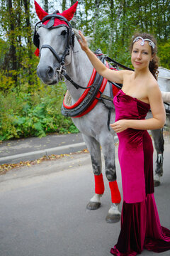 Pretty woman in red dress stands near horse in harness in green park