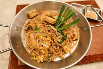 close up of prawn shrimp spring onion pad thai fried noodles in a steel pot dish and wooden tray. There is a bowl of salad and sauce on the side.