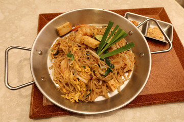 close up of prawn shrimp spring onion pad thai fried noodles in a steel pot dish and wooden tray. There is a bowl of salad and sauce on the side.