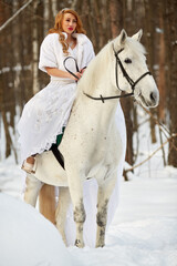 Young woman in white dress and white fur mantle on white horse in park