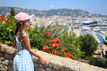 Girl standing near stone wall and red flowers and looks at Cannes, France