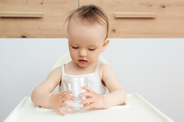 Sweet baby girl holding glass and drinking water. Happy baby sitting in a baby chair in the kitchen.