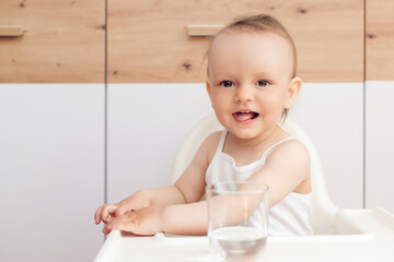 Sweet baby girl holding glass and drinking water. Happy baby sitting in a baby chair in the kitchen.