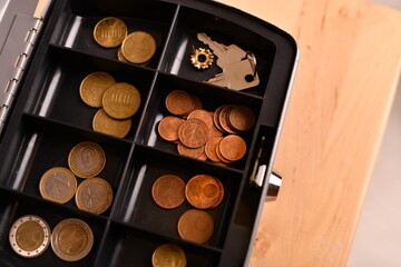 A black coin box filled with various coins, including copper and gold-toned currency, placed on a wooden surface. The image captures the details of the coins and the compartments of the box.
