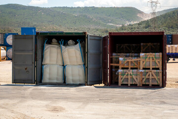 Two containers positioned side by side on the concrete floor, with the door open, one containing sacks and the other containing cases of processed marble.