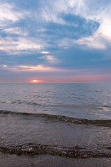 This photograph captures the quiet majesty of a sunset over the sea, with the sun dipping below the horizon, its light diffused by a veil of clouds. The sky is a canvas of soft blues and subtle pinks