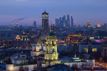 Bell tower of church of St. Sergius of Radonezh of Novospassky Monastery and high buildings in Moscow, Russia at evening