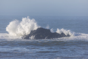A large rock is in the ocean with a wave crashing over it