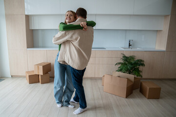Cheerful inspired husband and wife happy embrace among unpacked things, celebrating long-awaited...