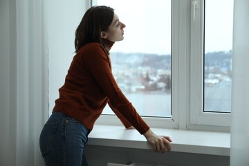 Sad young woman near window indoors, space for text