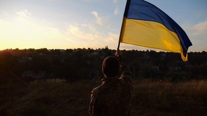 Young man in military uniform waving flag of Ukraine against beautiful sunset at background. Male ukrainian army soldier lifted national banner at countryside. Victory against russian aggression - 755758487