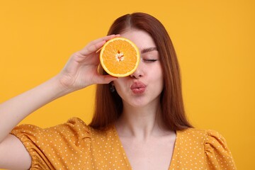 Beautiful woman covering eye with half of orange and sending air kiss on yellow background