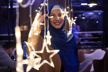 In a modern restaurant ambiance, a woman in a hijab captures a selfie beside glowing lights, showcasing contemporary style and cultural diversity in a trendy urban setting.
