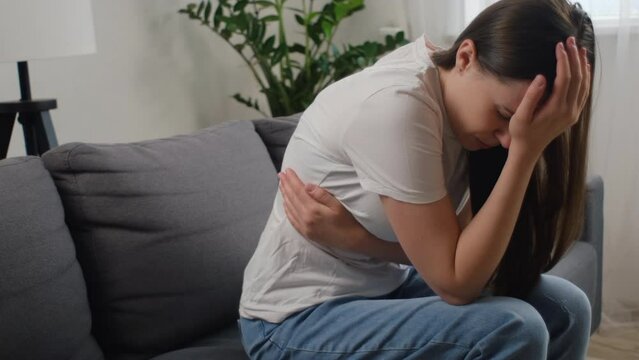 Unhealthy sad young woman suffering from menstrual pain. Girl with hands squeezing belly having painful stomach ache or period cramps sitting on couch. Abdominal pain and painful periods concept