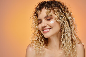 happy woman with wavy hair and cosmetic cream on face smiling with closed eyes on pastel backdrop