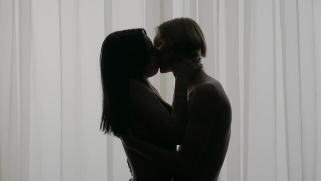 Young loving couple silhouettes in apartment. Man and woman half naked sensual lovers standing against window together, passion and intimacy kissing.