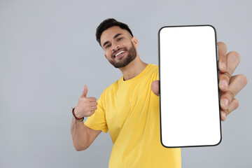 Happy man holding smartphone with empty screen and showing thumbs up on grey background, space for...