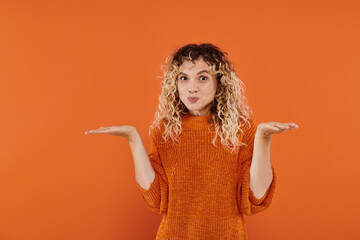 confused curly woman in knitted sweater puffing cheeks and showing shrug gesture on orange