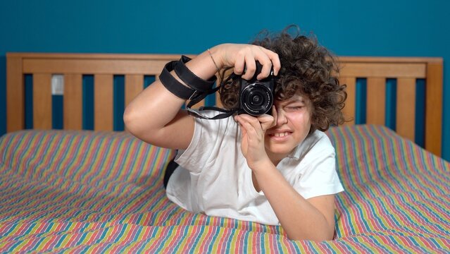 Funny and happy 9 year old boy in bed at home learns to take photographs with a camera - photography and lifestyle lessons in apartment during  childhood