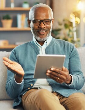 Cheerful senior man having video call on tablet sitting on the couch at home elderly man wearing eyeglasses staying in touch with friends and family using online video call connecting with people 