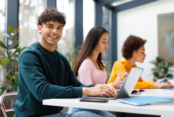 Cheerful latin male student with classmates using laptops