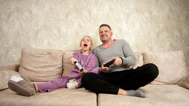 A father spends time with his little daughter, sitting on the sofa and playing a game console. Weekend activities, free time, home entertainment and video games concept.