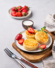 Cottage cheese, cheesecakes or syrniki with strawberries and mint leaves on a cutting board  on a light background with sour cream and napkin and cutlery.