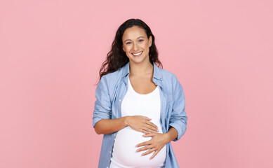 Cheerful expecting lady embracing big tummy, pink background