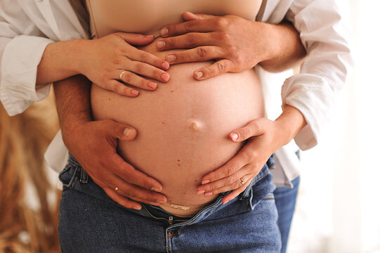 Close up photo of pregnant woman in white shirt and blue jeans and her husband holding their hands on pregnant belly. Horizontal picture. Family concept
