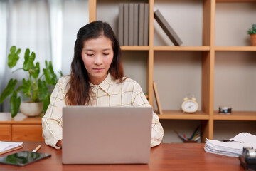 Asian adult female, engrossed in work, sitting at desk with laptop, tablet, papers; interior, daytime. Concentrated young office worker reviews documents, digital devices on wooden table, calm setting