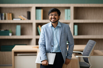 Happy Indian businessman stands near his office desk holding laptop