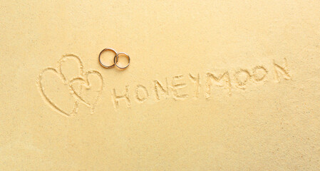 Word Honeymoon written on sand, hearts two golden rings, top view