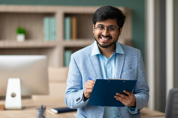 Professional Indian HR manager guy holds clipboard at modern workplace