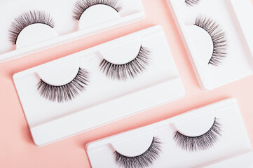 Different fake eyelashes in boxes on trendy pastel pink background. Makeup accessories and beauty cosmetics products for women. Top view, flat lay.