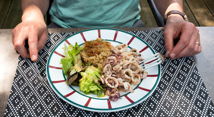 Man preparing to eat his meal at traditional French Basque country restaurant. Squid rings, rice with ratatouille, fresh green salad aesthetically served on plate with national Basque colors.